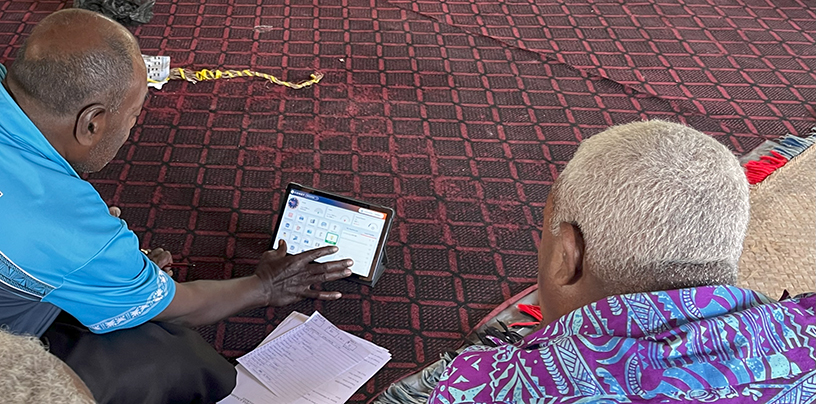 Two men seen from behind sitting on a red mat. One touches a touchscreen (iPad).