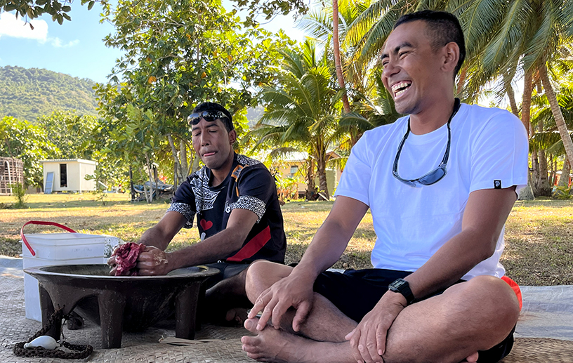 Two men sit in traditional style on the ground. One laughs while the other prepares kava in a tanoa (wooden bowl).