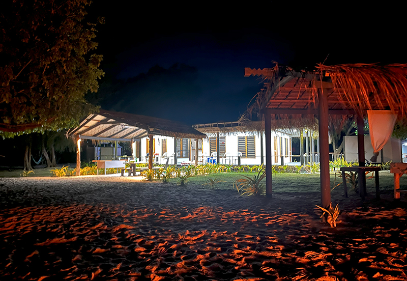 A one story house and a ramada with thatched roofs at night set against the glow of a fire burning out of frame.