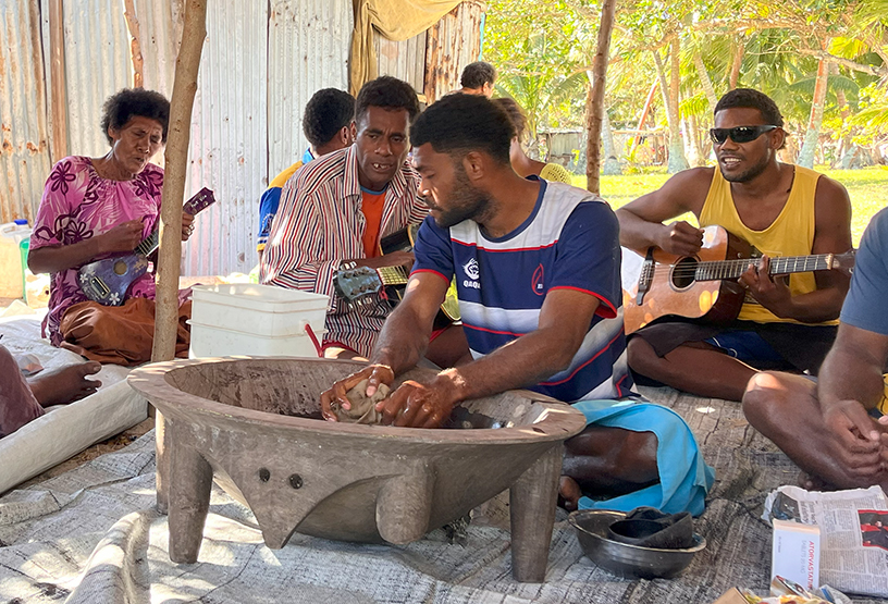 A group of people sitting on a mat on the ground. Two play guitar. Most sing. One prepares kava in a wooden bowl.