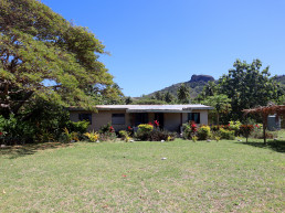 A one-story house in Marou Village Fiji with the mountain rising in the background and a large shade tree to the left.