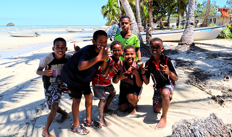 Six children kneeling and giving thumbs up signs on the beach.