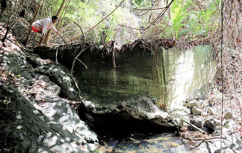 A triangular wall of poured-in-place concrete rests on the banks of a creek. A shadow shows were the water has found a path to flow beneath the wall. A person crouches on top of the wall on the left side of the frame pointing to the right.