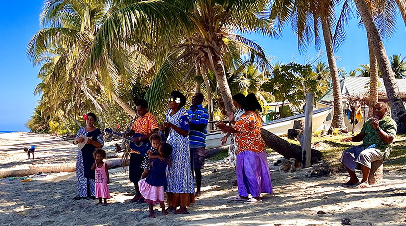 Villagers singing and playing guitar on the beach under coconut palms, dressed in colorful clothing.