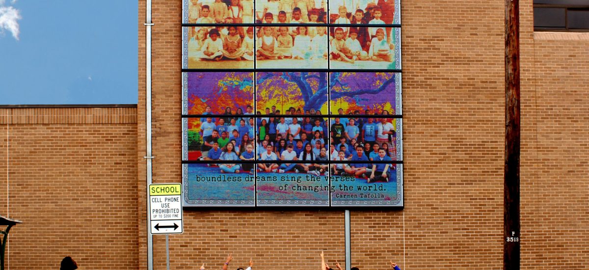 En-Aquellos-Tiempos-Photo-w-Students Students pose in front of the Land Art Generator Solar Mural Installation on the JT Brackenridge Elementary School in San Antonio, TX that features their class below an image of the class of 1906, showing the continuity of life in the neighborhood over more than a century. Artwork by Adriana Garcia, Art Direction by Penelope Boyer, Poetry by Carmen Tafolla, Photography on artwork by Antonia Padilla. Photo by Imgard Rop. Project partners include: Esperanza Peace & Justice Center, Land Art Generator, Mr. Vega’s 5th grade class at JT Brackenridge, Community elders from the Westside, South Texas Solar Systems, Mission Solar Energy, Outback Power, Sistine Solar, and Unirac.