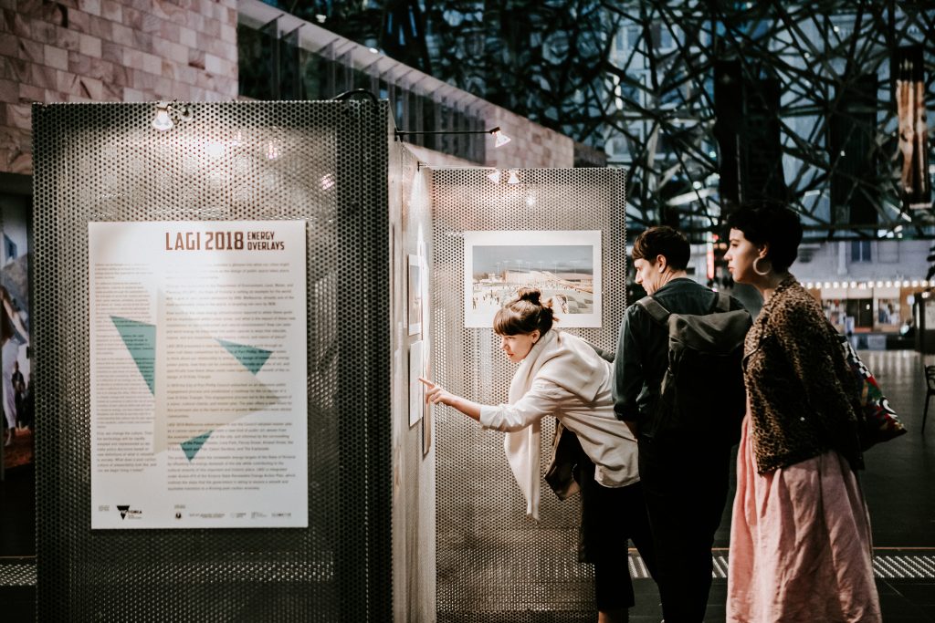 Visitor to the LAGI 2018 Melbourne exhibition leans over to read one of the display boards during the award ceremony, exhibition, and book launch at Fed Square. She points at the board while two other people stand behind also  looking. It is evening and the lights have come on in the street in the background. It is a large space with complex ceiling structure and with lighting focused on the art boards.
