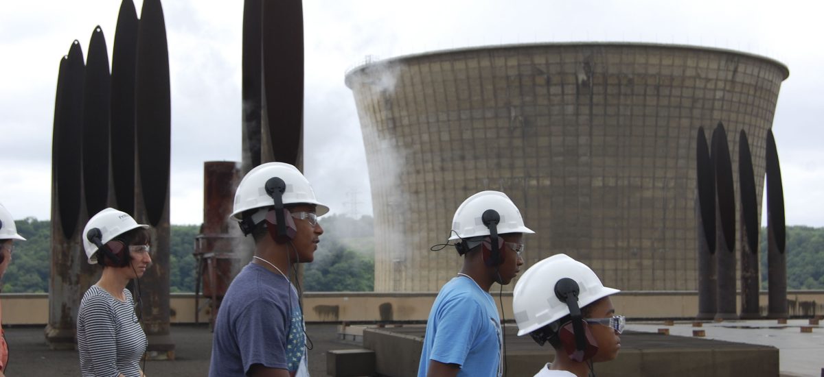 day3_1977 Youth tour sites around Pennsylvania to get an understanding of the energy mix of Western PA, during the 2015 Art+Energy Camp. In this photo we see Elizabeth Monoian (left) and three Art + Energy Camp participants wearing hard hats and walking on the roof of a coal fired power plant . The vent stacks and cooling tower can be seen behind them. The sky is grey.