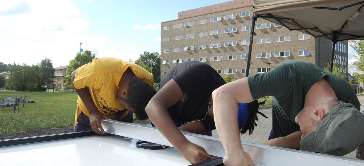 week6_00972 Youth help to construct a solar artwork during the 2015 Art+Energy Camp. In this photo we see Robert Ferry and two young Art + Energy Camp participants tightening screws on the frame of an upside down solar module prior to installing it on the artwork.