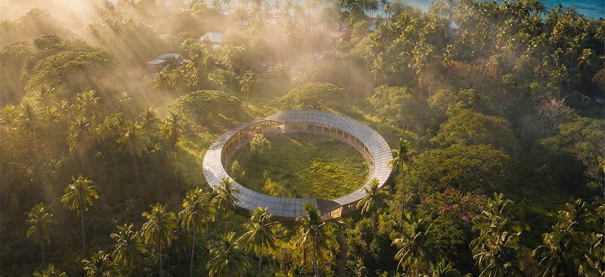 The O_Aerial beams of sunlight illuminate a ring-shaped pavilion clad in solar photovoltaic modules. The aerial view looks down on a Fijian coastal village adjacent to The O installation.